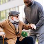 A man helping an older gentleman walk with his cane.